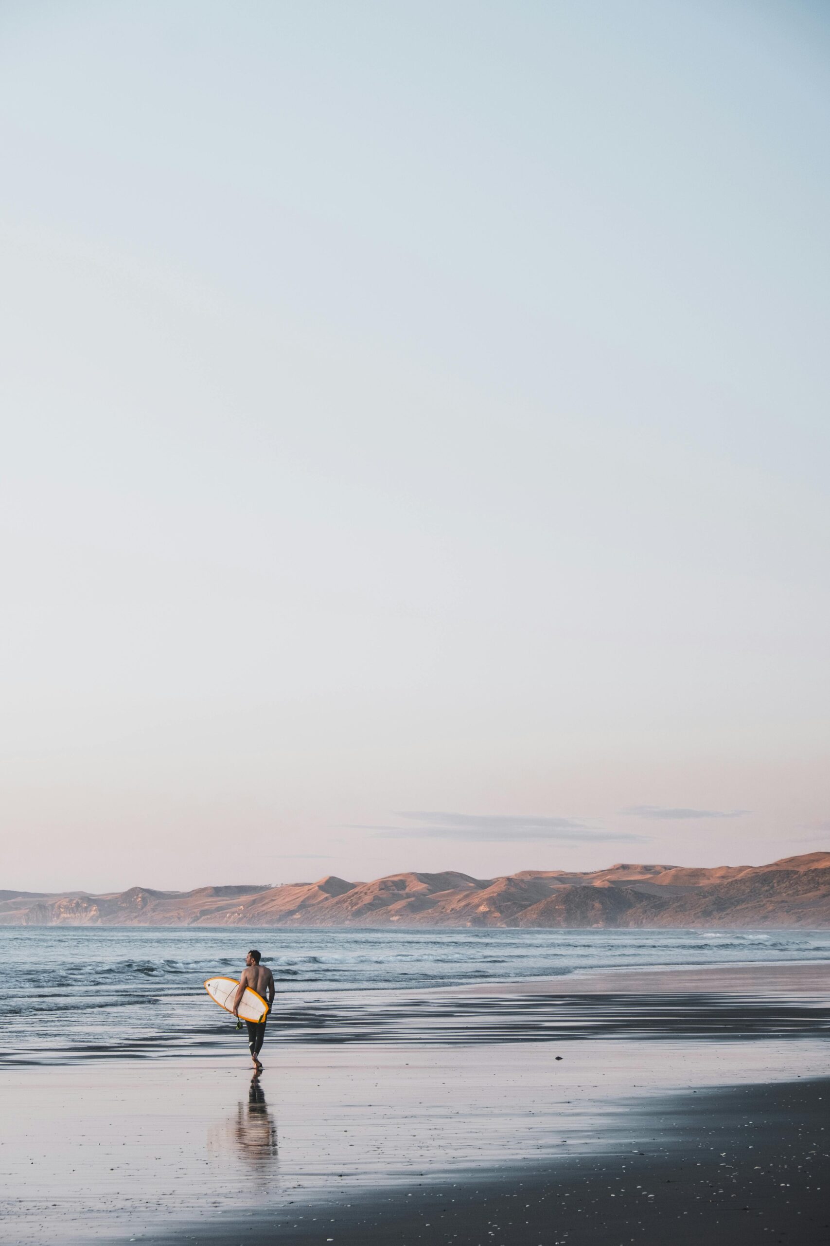 pexels-photo-4007605-4007605 A lone surfer with a surfboard walks along the serene beach at sunset in New Zealand.
