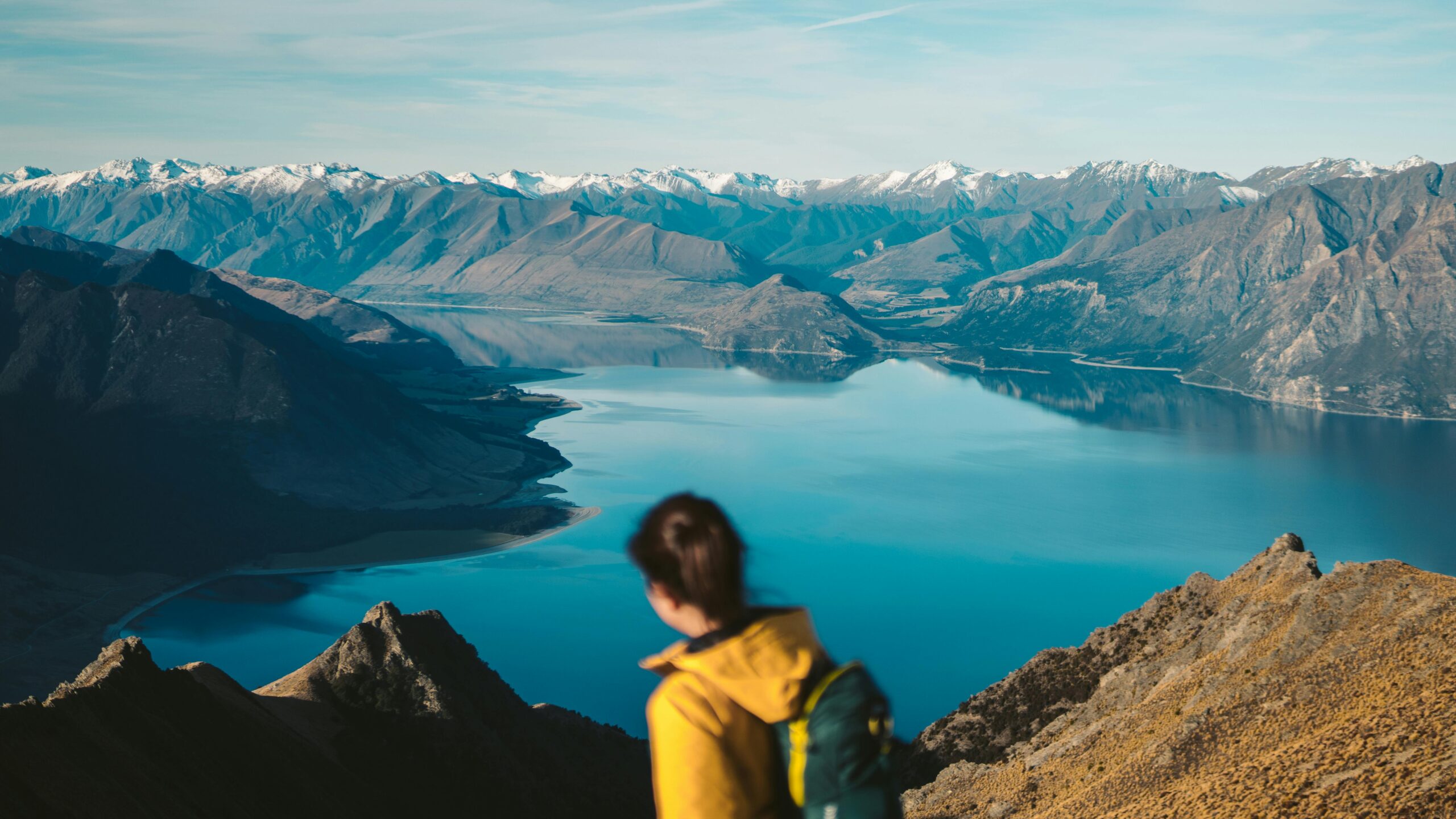 A woman hiker enjoys a picturesque view of a tranquil lake in Otago, New Zealand, with snow-capped mountains.