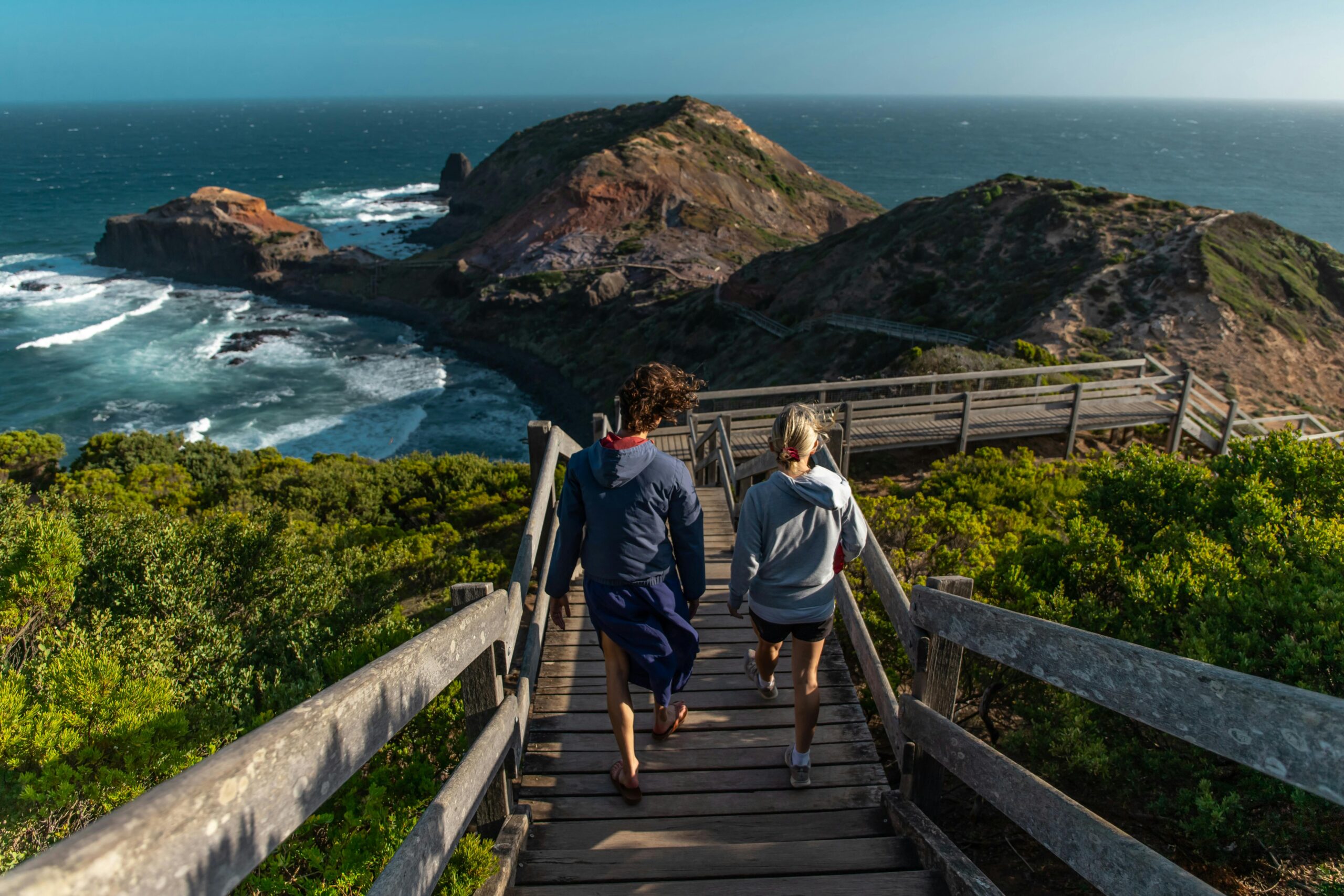 Two people walking down wooden stairs towards a scenic coastal view in Melbourne, Australia.