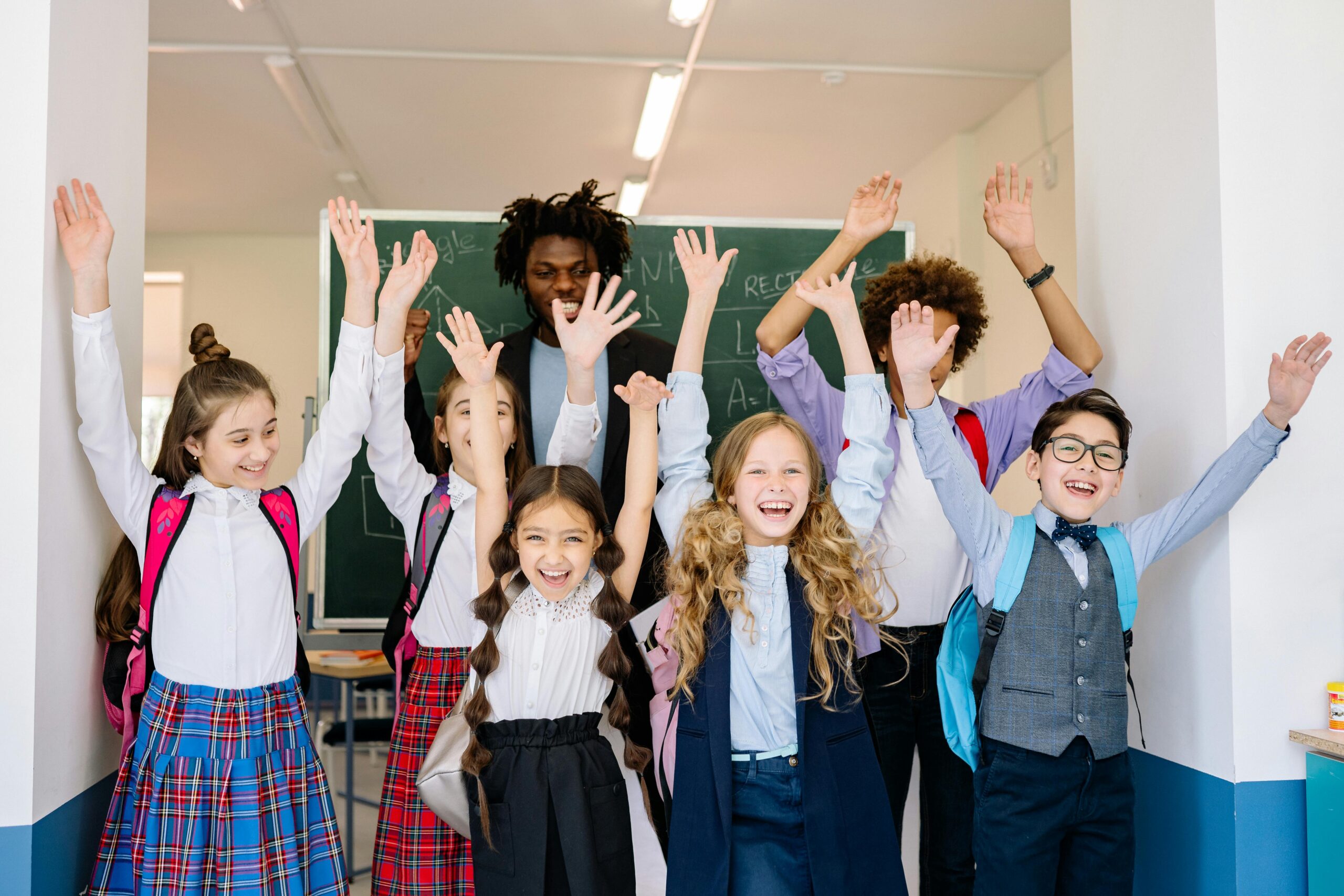 Joyful group of diverse school children raising their hands in an indoor classroom setting.